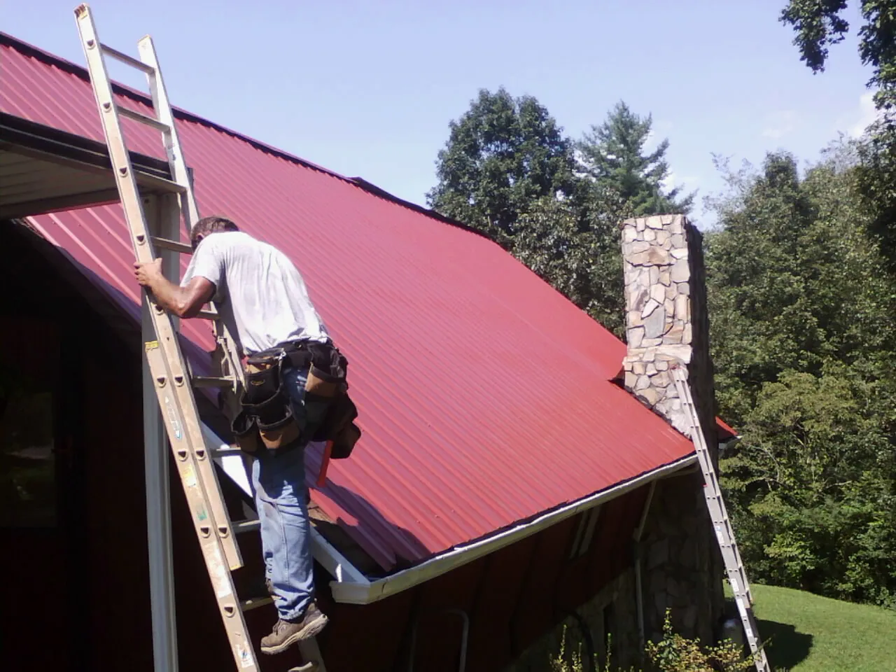 Roofer on a ladder beside a red metal roof and stone chimney — Soffit & Fascia Repair by Roofing Brothers in Effingham